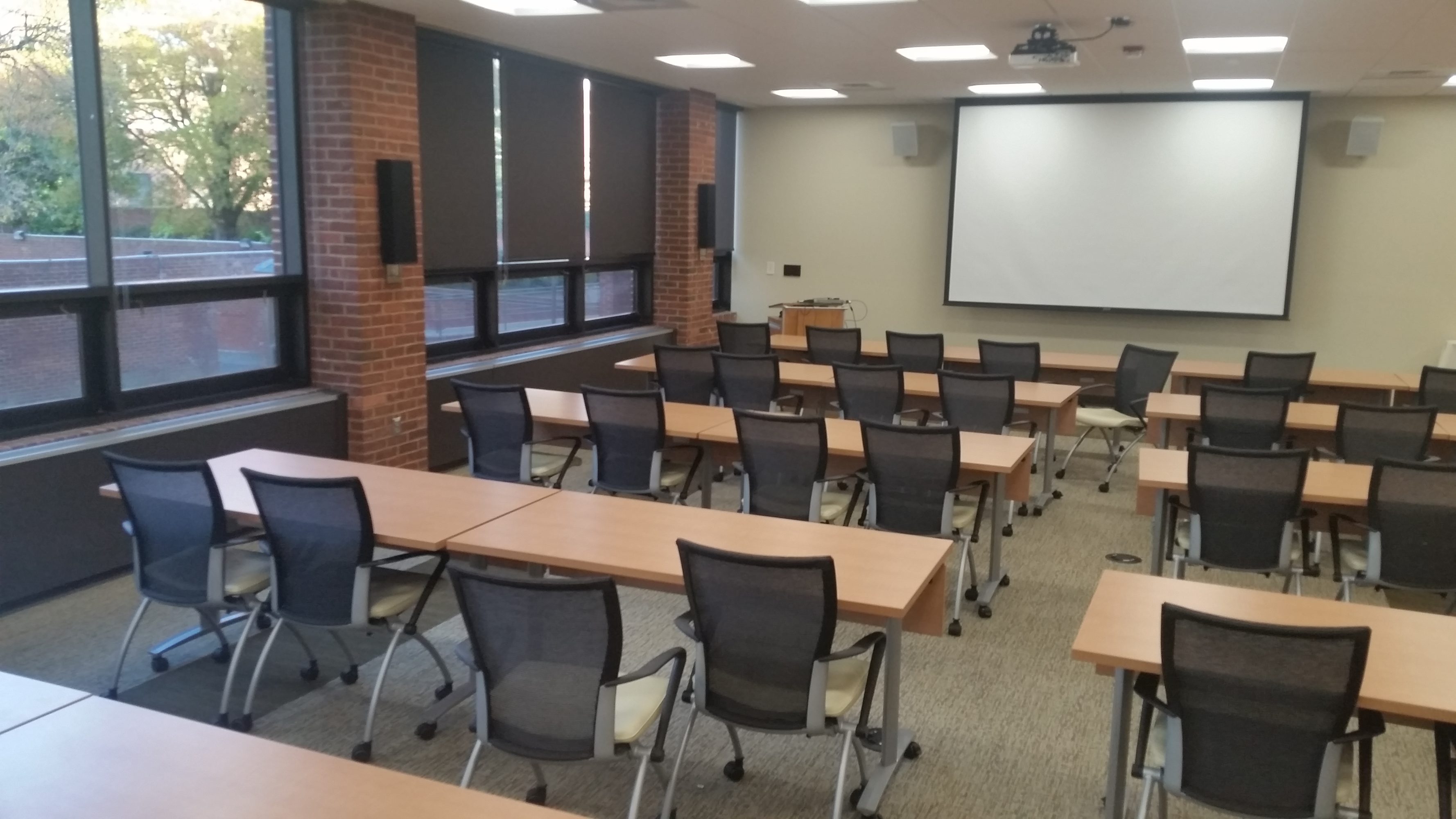 An empty meeting room with rolling chairs at desks facing the front with a screen